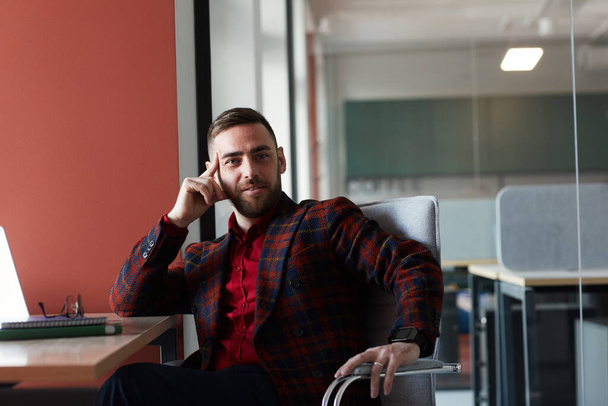Retrato de homem de negócios barbudo elegante olhando para a câmera e sorrindo enquanto sentado na mesa e posando contra a parede vermelha no escritório, espaço de cópia - Foto, Imagem
