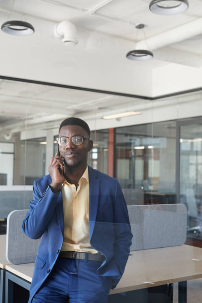 Vertical portrait of successful African-American businessman speaking by smartphone while leaning on desk in modern office interior, copy space - 写真・画像