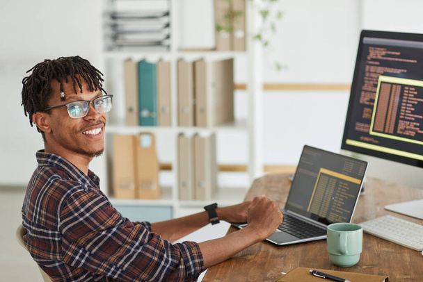 Portret uśmiechniętego afro-amerykańskiego dewelopera IT patrzącego w kamerę podczas pisania na klawiaturze czarno-pomarańczowego kodu programowania na ekranie komputera i laptopie, przestrzeń do kopiowania - Zdjęcie, obraz