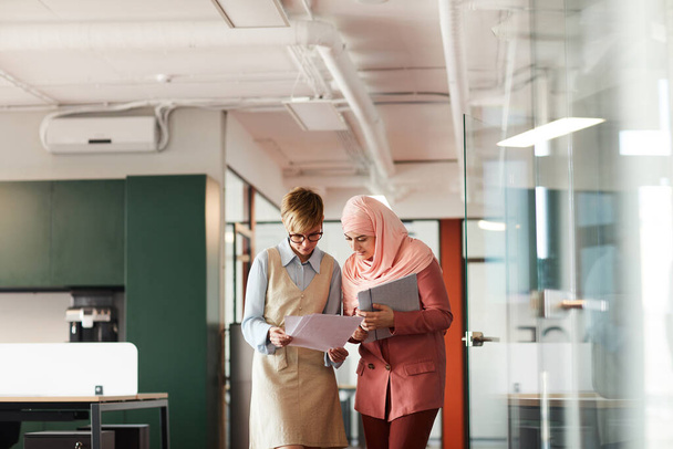 Širokoúhlý portrét manažerky hovořící s mladou muslimskou podnikatelkou a držící dokumenty při chůzi směrem ke kameře v interiéru kanceláře, kopírovací prostor - Fotografie, Obrázek
