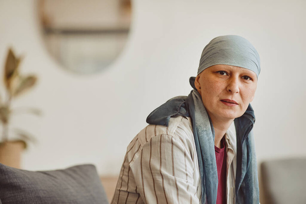 Θερμαινόμενο πορτραίτο φαλακρής ενήλικης γυναίκας που φορά μαντίλα κοιτάζοντας την κάμερα ενώ ποζάρει σε minimal εσωτερικό χώρο, αλωπεκία και ευαισθητοποίηση σε θέματα καρκίνου, αντιγραφή χώρου - Φωτογραφία, εικόνα