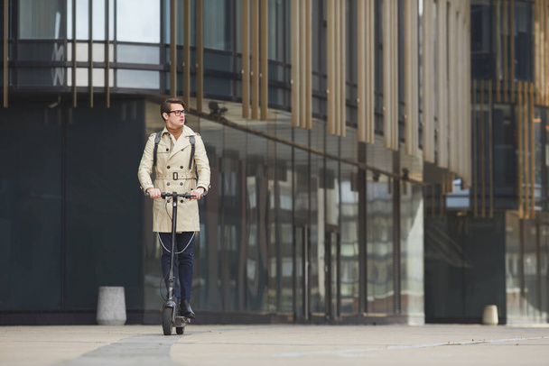 Széles látószögű portré a modern fiatal üzletember lovaglás elektromos robogó felé kamera, miközben ingázik dolgozni városi épületek a háttérben, másolás tér - Fotó, kép