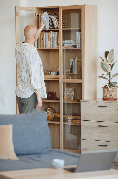 Vertical back view at bald person putting book on shelf on wooden bookcase in modern home interior, alopecia and cancer awareness, copy space - Fotó, kép