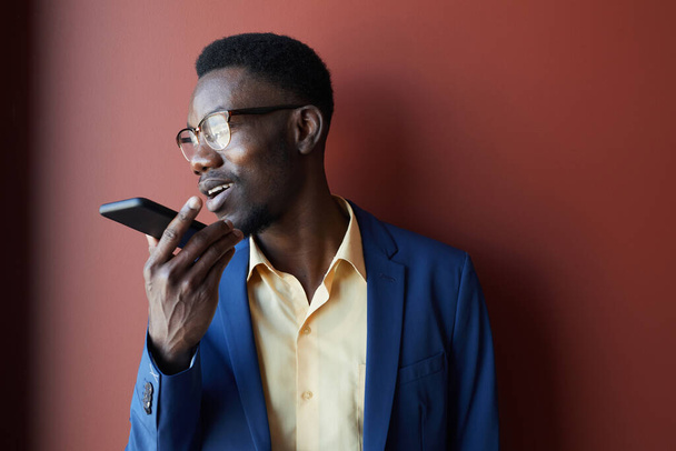 Portrait of elegant African-American man recording voice message via smartphone and wearing glasses while posing against maroon background, copy space - Фото, изображение