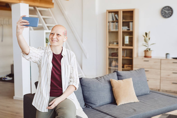 Minimal portrait of bald adult woman with head tattoo taking selfie and smiling happily while standing in modern home interior, alopecia and cancer awareness, copy space - Photo, Image