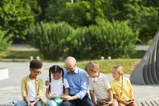 Vista frontal en el grupo multiétnico de niños usando tabletas y teléfonos inteligentes mientras están sentados en la fila al aire libre a la luz del sol con el hombre maduro ayudándoles, espacio para copiar - Foto, Imagen