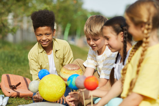 Multi-ethnic group of children sitting on green grass and holding model planets while enjoying outdoor astronomy lesson in sunlight, copy space - Photo, Image