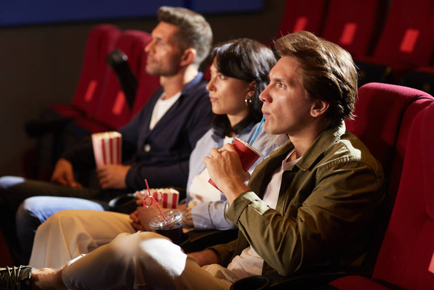 Retrato de vista lateral de jóvenes viendo películas en el cine mientras están sentados en fila en sillas de terciopelo rojo, se centran en el hombre bebiendo refresco a través de paja con expresión de cara tensa, espacio para copiar - Foto, Imagen