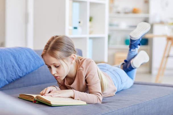 Full length portrait of cute blonde girl reading book or studying while lying on couch in cozy home interior, copy space - Photo, image