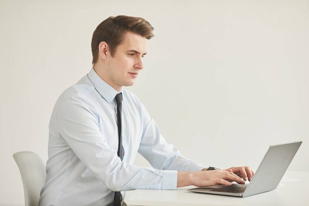 Minimal side view πορτρέτο του επιτυχημένου νεαρού επιχειρηματία που χρησιμοποιεί φορητό υπολογιστή, ενώ κάθεται στο γραφείο και εργάζεται στο γραφείο κατά λευκό τοίχο, αντιγραφή χώρου - Φωτογραφία, εικόνα
