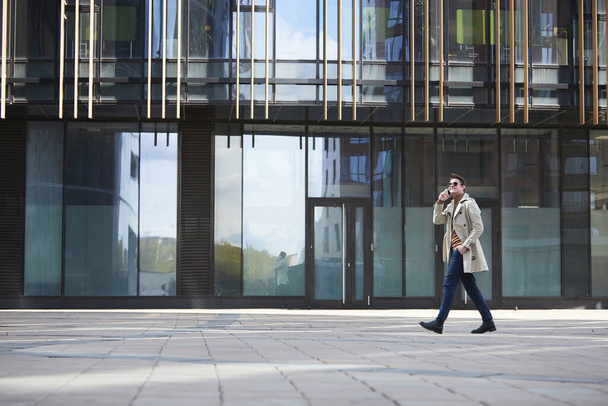 Retrato de gran angular del elegante hombre de negocios que lleva gabardina caminando a través de disparo mientras habla por teléfono inteligente, edificio urbano de vidrio en el fondo - Foto, imagen