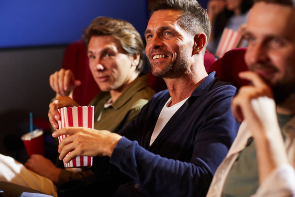 Группа улыбающихся мужчин, которые смотрят кино в кинотеатре и едят попкорн, сидя в ряд на красных сиденьях, копируют пространство - Фото, изображение