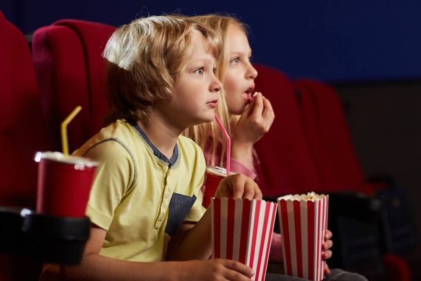 Widok z boku portret dwójki dzieci z otwartymi ustami oglądających film w kinie i jedzących popcorn, przestrzeń kopiująca - Zdjęcie, obraz