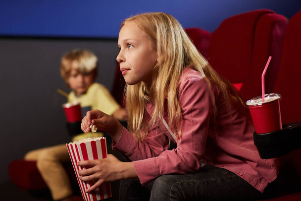 Side view portrait of blonde teenage girl watching movie in cinema theater and eating popcorn, copy space - Foto, Bild