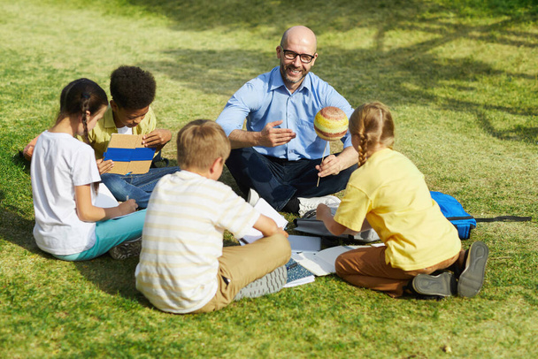Высокоугловой портрет лысого учителя, который указывает на модель планеты и улыбается, разговаривая с группой детей во время занятий на открытом воздухе под солнечным светом, копирует пространство - Фото, изображение