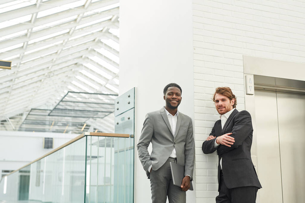 Széles látószögű portré két sikeres üzletemberről, akik öltönyt viselnek és mosolyognak a kamera előtt, miközben irodaépület belsejében állnak, másolja le a helyet - Fotó, kép