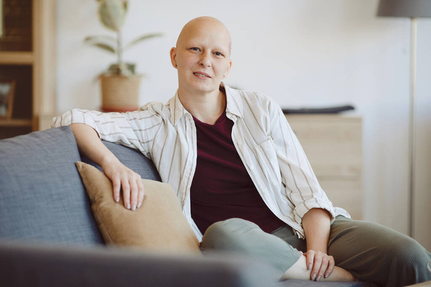 Retrato de mulher adulta careca olhando para a câmera enquanto sentado no sofá no interior da casa moderna, alopecia e consciência do câncer, espaço de cópia
 - Foto, Imagem