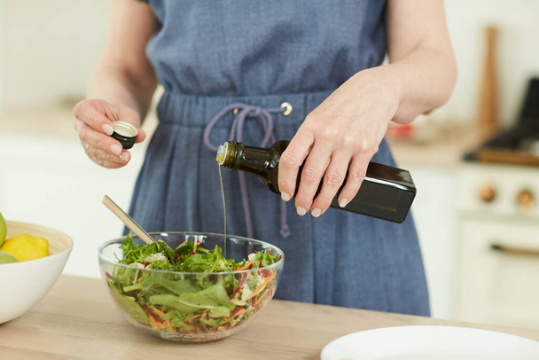 Primer plano de la mujer elegante irreconocible la adición de aceite de oliva a la ensalada saludable en un tazón de vidrio mientras disfruta de la cocina en casa, espacio para copiar - Foto, imagen