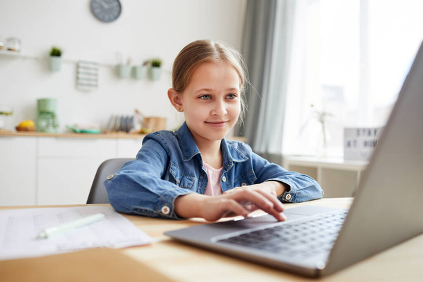 Портрет милой маленькой девочки с помощью ноутбука и улыбаясь во время учебы в Интернете в уютном интерьере, копировать пространство - Фото, изображение