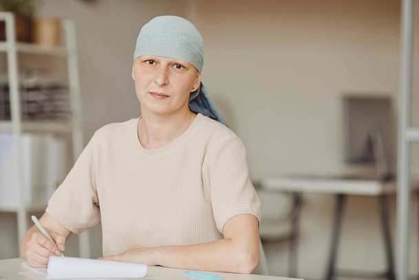 Portrait taille chaude de femme chauve mature portant un foulard regardant la caméra tout en remplissant des papiers dans le bureau des médecins, l'alopécie et la sensibilisation au cancer, copier l'espace - Photo, image