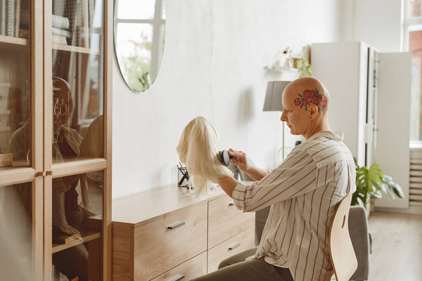 Boční pohled portrét moderní plešaté ženy kartáčování paruky při sezení u zrcadla v domácím interiéru, alopecie a povědomí o rakovině, kopírovat prostor - Fotografie, Obrázek