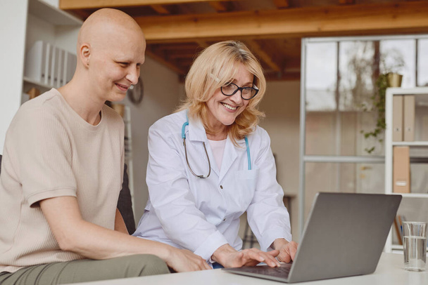 Meleg tónusú portré egy vidám női orvosról, aki laptopot használ és kopasz beteggel beszél az alopéciáról és a rák gyógyulásáról szóló konzultáció során, másolás - Fotó, kép