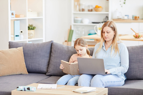 Portrait of adult working mother using laptop while sitting on couch at home with cute daughter reading book beside her, copy space - Photo, Image