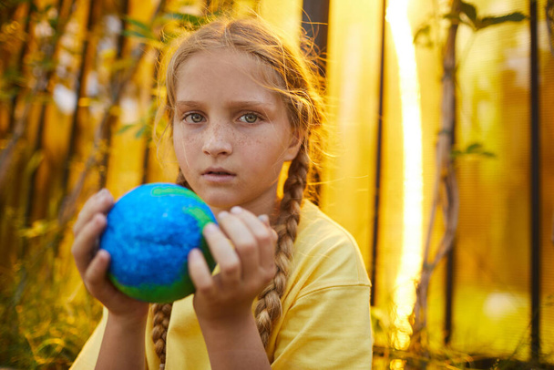 Ritratto di ragazzina lentigginosa che tiene il pianeta in mano e guarda la macchina fotografica mentre si trova all'aperto alla luce del sole, copia lo spazio - Foto, immagini