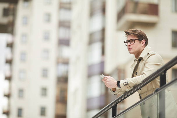 Grafische lage hoek portret van moderne jonge zakenman dragen trenchcoat en het houden van smartphone terwijl leunend op leuning buiten in stedelijke omgeving, kopiëren ruimte - Foto, afbeelding