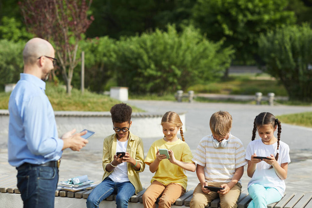 Multiethnische Gruppe von Kindern, die digitale Tablets verwenden, während sie während des Unterrichts im Freien mit einem lächelnden männlichen Lehrer in einer Reihe auf einer Bank sitzen, Kopierraum - Foto, Bild