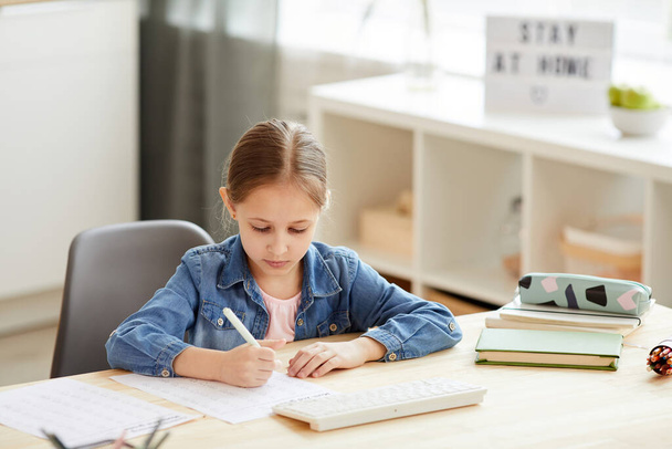 Warm getönte Porträt des netten kleinen Mädchens, das Hausaufgaben am Schreibtisch macht, während es zu Hause in gemütlicher Inneneinrichtung studiert, Kopierraum - Foto, Bild