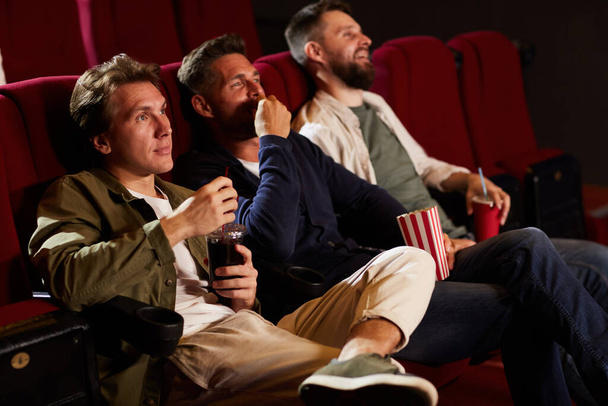 Группа друзей-мужчин смотрит кино в кинотеатре и ест попкорн, сидя в ряд на красных сиденьях, скопировать пространство - Фото, изображение