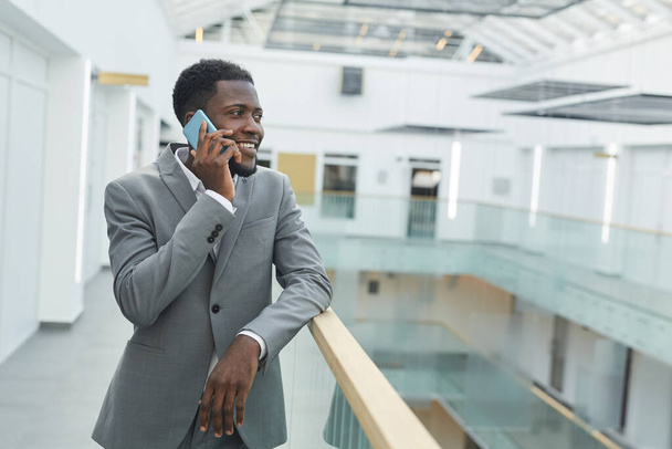 Taille omhoog portret van succesvolle Afro-Amerikaanse zakenman spreken per smartphone en glimlachen terwijl leunend op balkon reling in modern kantoorgebouw interieur, kopieer ruimte - Foto, afbeelding