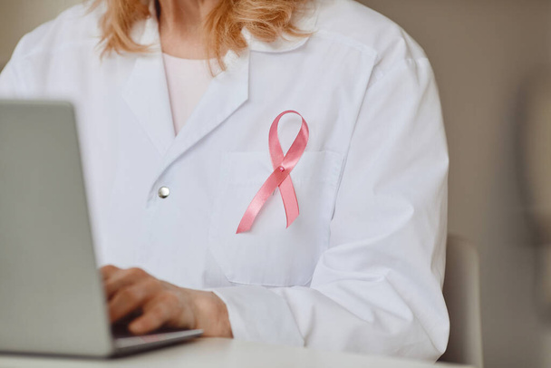 Close up background of pink ribbon pinned on white lab coat of female doctor using laptop, breast cancer awareness symbol, copy space - Photo, Image