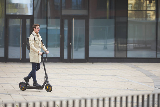 Wide angle portrait of modern young businessman riding electric scooter while commuting to work with urban city buildings in background, copy space - Photo, Image