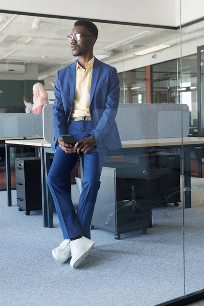 Vertikální celovečerní portrét úspěšného afroamerického podnikatele, jak odvrací pohled a opírá se o stůl v moderním kancelářském interiéru, kopíruje prostor - Fotografie, Obrázek
