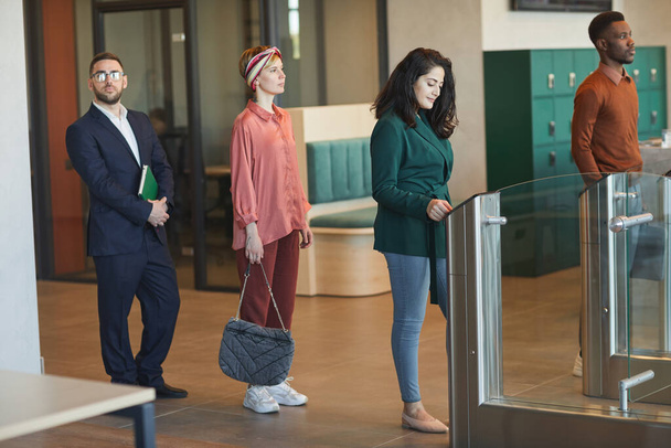 Сторона зору на багатоетнічну групу людей, що стоять у черзі, переходячи через автоматизовані ворота, щоб увійти в офісну будівлю, зосередитися на молодих близькосхідних жінок, які розмахують ідентифікаційними картками, копіювальний простір - Фото, зображення