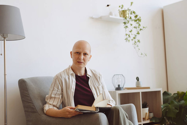 Ritratto di donna matura calva che guarda la macchina fotografica mentre legge il libro seduta in comoda poltrona a casa, alopecia e consapevolezza del cancro, spazio copia - Foto, immagini