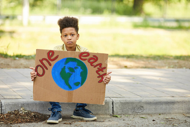 Pełna długość portret Afroamerykanina chłopca trzymając znak z ONE EARTH i patrząc w obiektyw podczas protestu o przyrodę na zewnątrz, kopiuj przestrzeń - Zdjęcie, obraz