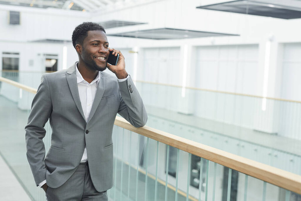 Cintura-se retrato de empresário confiante afro-americano falando por smartphone e sorrindo enquanto se inclina no corrimão varanda no interior do edifício de escritório moderno, espaço de cópia - Foto, Imagem