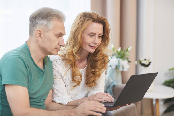 Side view portrait of modern mature couple using laptop together while sitting on couch in home interior, copy space - Foto, Imagem