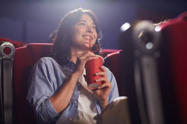 Ritratto ad angolo basso di giovane donna sorridente nel cinema che guarda il film da sola e beve soda, guarda lo schermo, copia lo spazio - Foto, immagini
