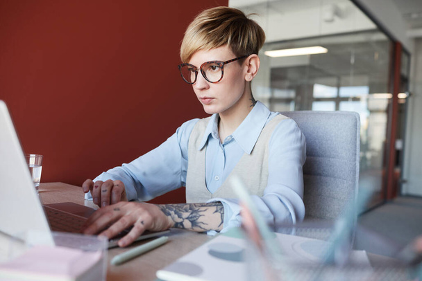 Portret van een moderne getatoeëerde zakenvrouw met behulp van laptop aan het bureau tijdens het werken in het kantoor tegen de rode muur, kopieer ruimte - Foto, afbeelding