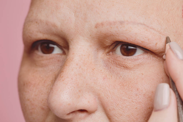 Extremo close-up de sardento careca mulher desenho sobrancelhas e fazendo maquiagem contra fundo rosa no estúdio, alopecia e consciência do câncer, espaço de cópia
 - Foto, Imagem