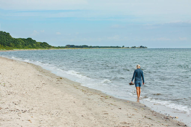 Young blonde woman walking in the water on a beach by the sea with her shoes in her hand. Woman seen from behind fron a distance. Waves rolling in at the shore. - Fotoğraf, Görsel