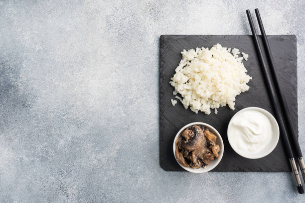Boiled white rice and mushrooms in sour cream on a black stand with chopsticks. Grey concrete background Copy space - Foto, immagini