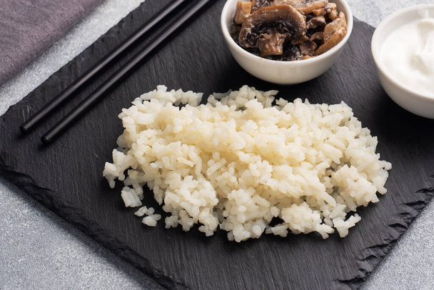 Boiled white rice and mushrooms in sour cream on a black stand with chopsticks. Grey concrete background close up. - Foto, immagini