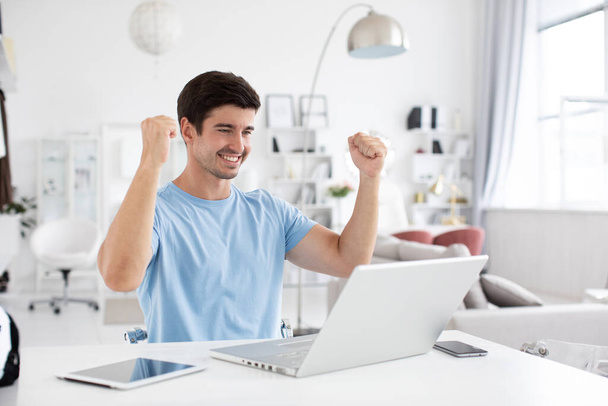 Handsome young man sitting at his desk, celebrating success with arms raised while looking at his laptop screen. Distance job. - Foto, imagen