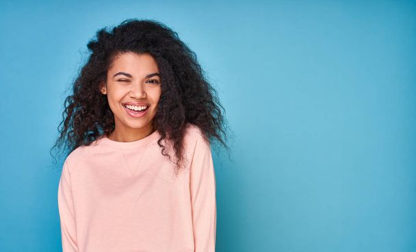 Positive dark-skinned curly woman winks, feels cheerful and cheerful, wears a beige long-sleeved sweater, isolated on blue studio background. Copy space. - Fotó, kép