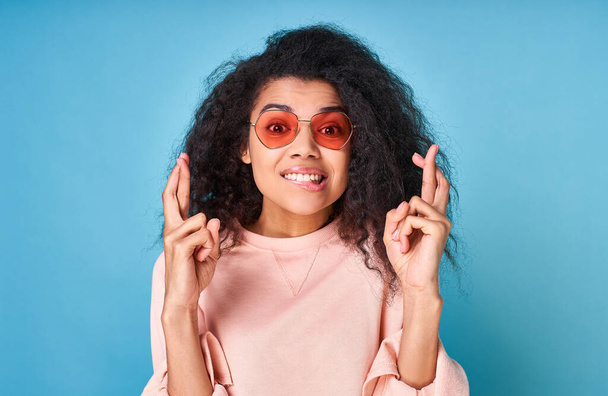 A positive black woman with an afro hairstyle, in red sun glasses, looks straight, makes a prayer gesture, smiles broadly, hopes that dreams come true, isolated on a blue background. - Foto, Imagem